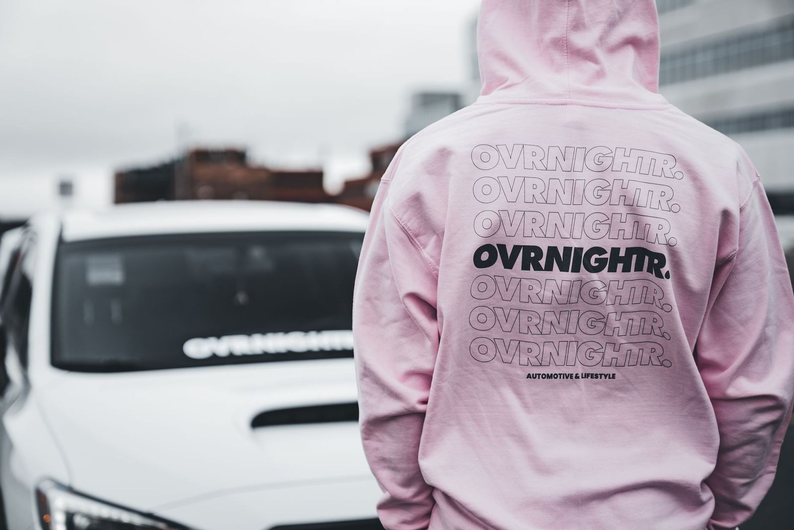 Stylish back view of person in pink hoodie standing near white sports car in urban setting.