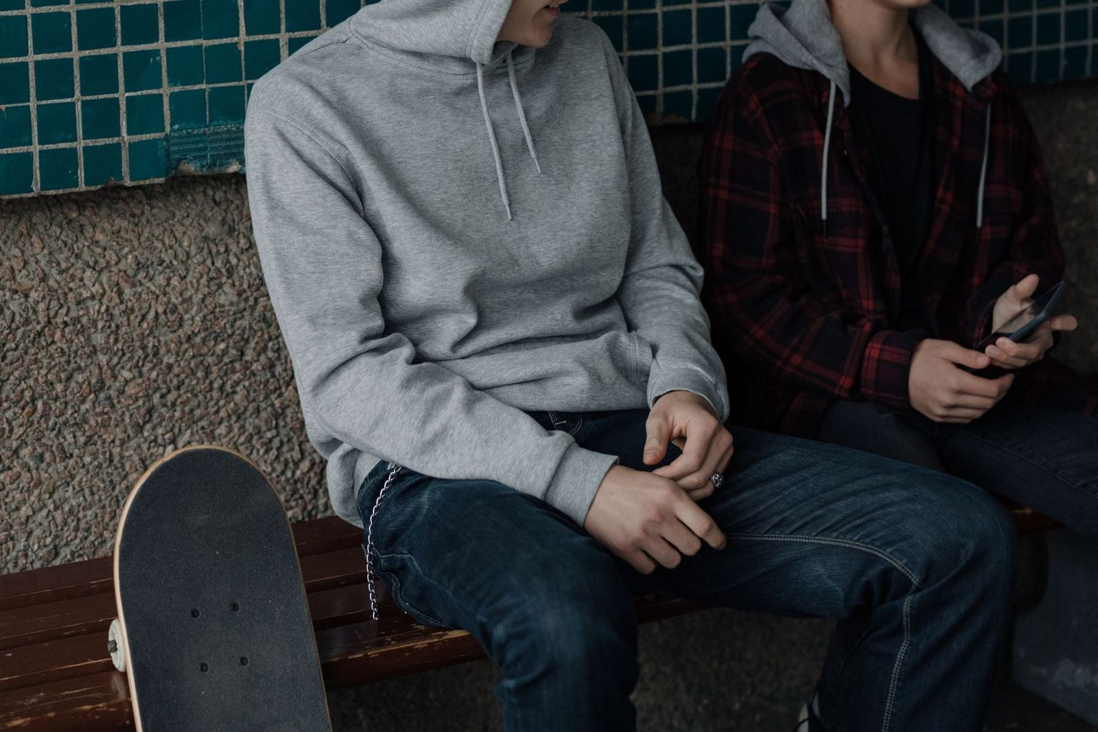 Two teenagers sitting on a bench with a skateboard, wearing casual hoodies in an urban environment.
